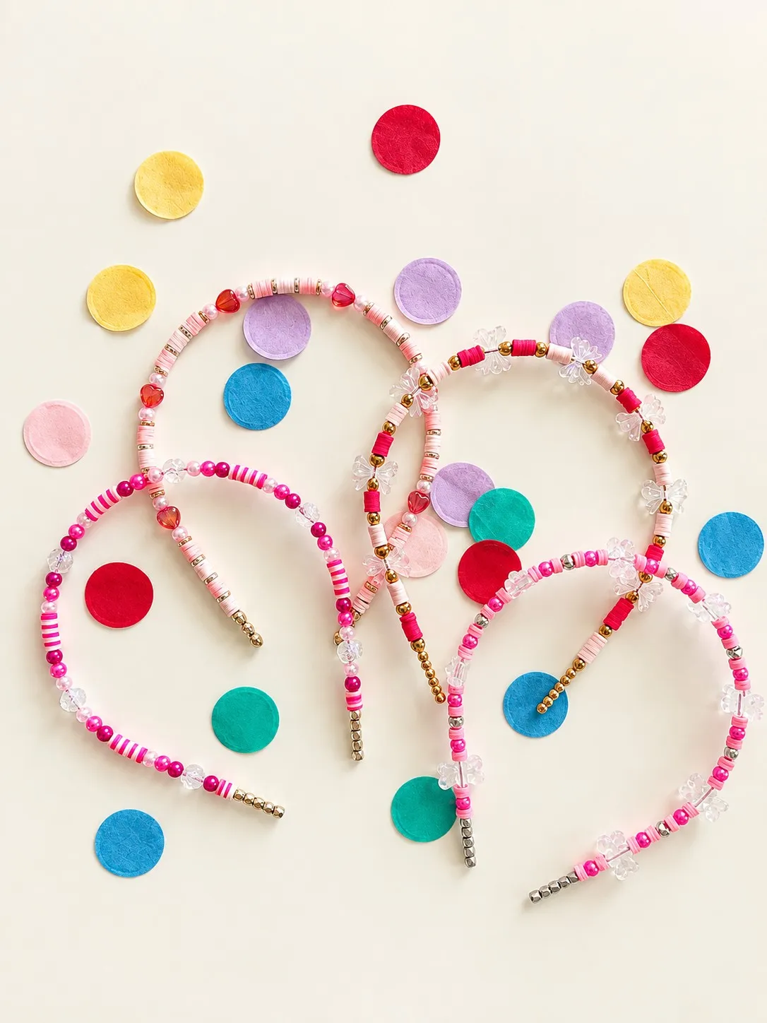 Four pink beaded headbands with red heart beads and clear bow charms, crossing over scattered paper confetti.